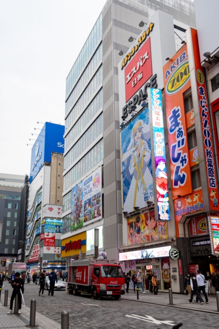 Colorful anime billboards and electronic store signs in Akihabara, Tokyo’s famous electronics and otaku district, with people walking and a Coca-Cola truck passing by.