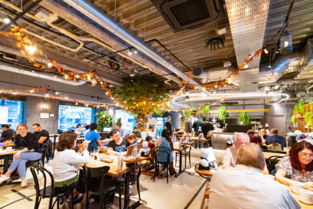 A lively scene inside Dawn Café in Tokyo, where customers sit at tables alongside small humanoid robots assisting with service. The modern interior is decorated with hanging greenery and autumn leaves, creating a cozy yet futuristic atmosphere.