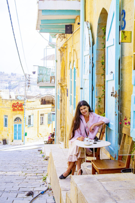 Woman with long dark hair in a pink dress enjoying coffee on a sunlit stone street with turquoise shutters.