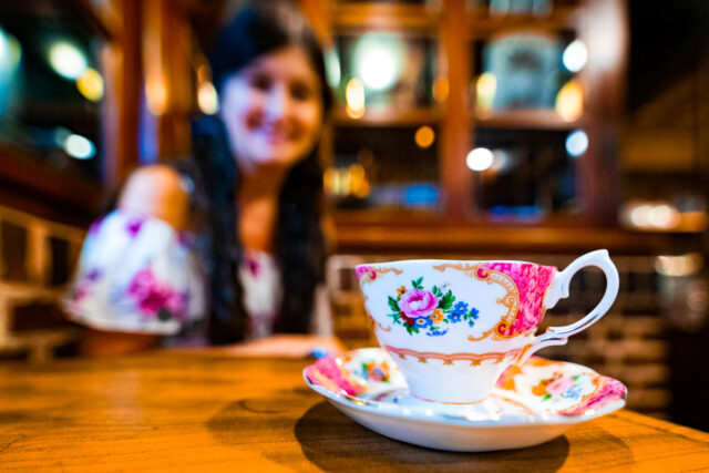 A floral porcelain teacup and saucer at Kurashiki Coffee Kan, with a blurred background of a woman with long hair sitting in the retro-style café.