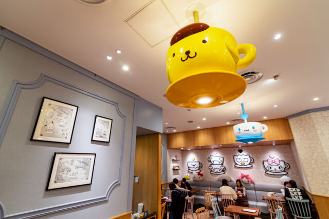Interior of the Sanrio Café in Ikebukuro, featuring ceiling lights shaped like Pompompurin and Cinnamoroll teacups, framed Sanrio sketches on the wall, and customers dining in a cozy, character-themed space.