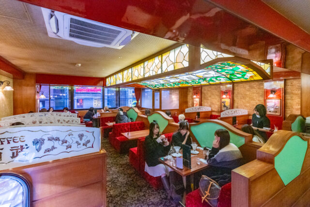 Retro-style kissaten café inside Seibu Shinjuku, featuring wooden booths with green accents, stained-glass ceiling panels, and red velvet seating. Customers enjoy coffee and desserts in a cozy, nostalgic atmosphere while a waitress serves a table.