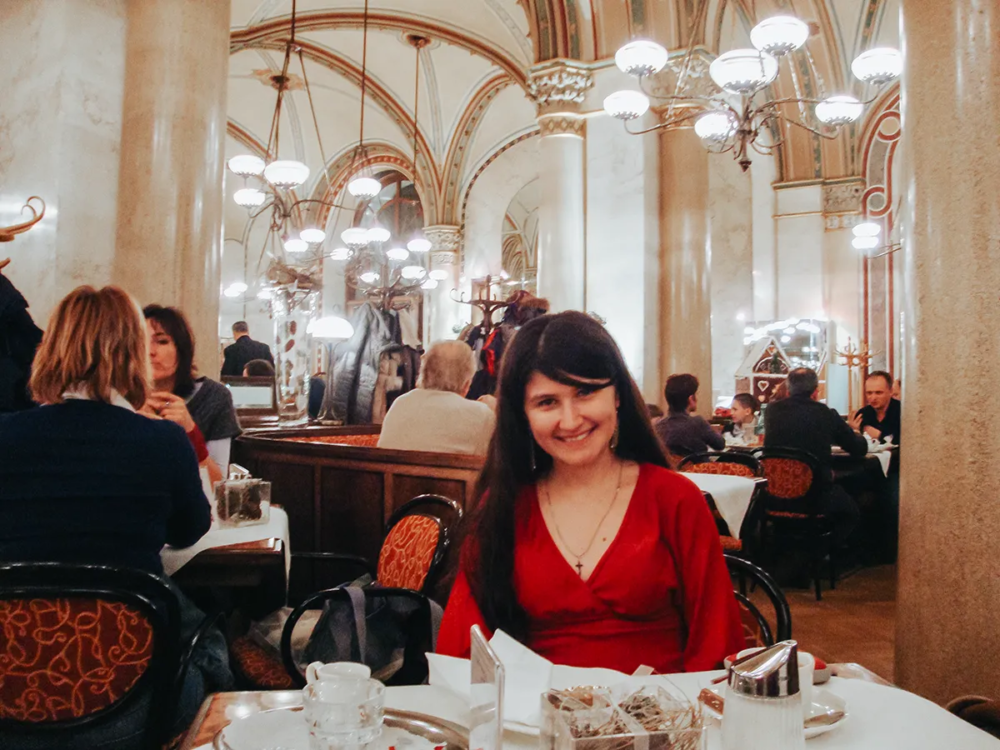Ket in red dress sitting at the Café Central in Vienna with vaulted ceilings, chandeliers, and guests enjoying the historic coffeehouse atmosphere