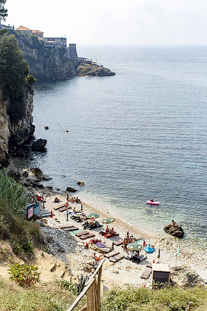 Aerial view of a small pebble beach in Ulcinj's Liman area with sunbathers and loungers, rocky cliffs and the Adriatic Sea in the background, Montenegro