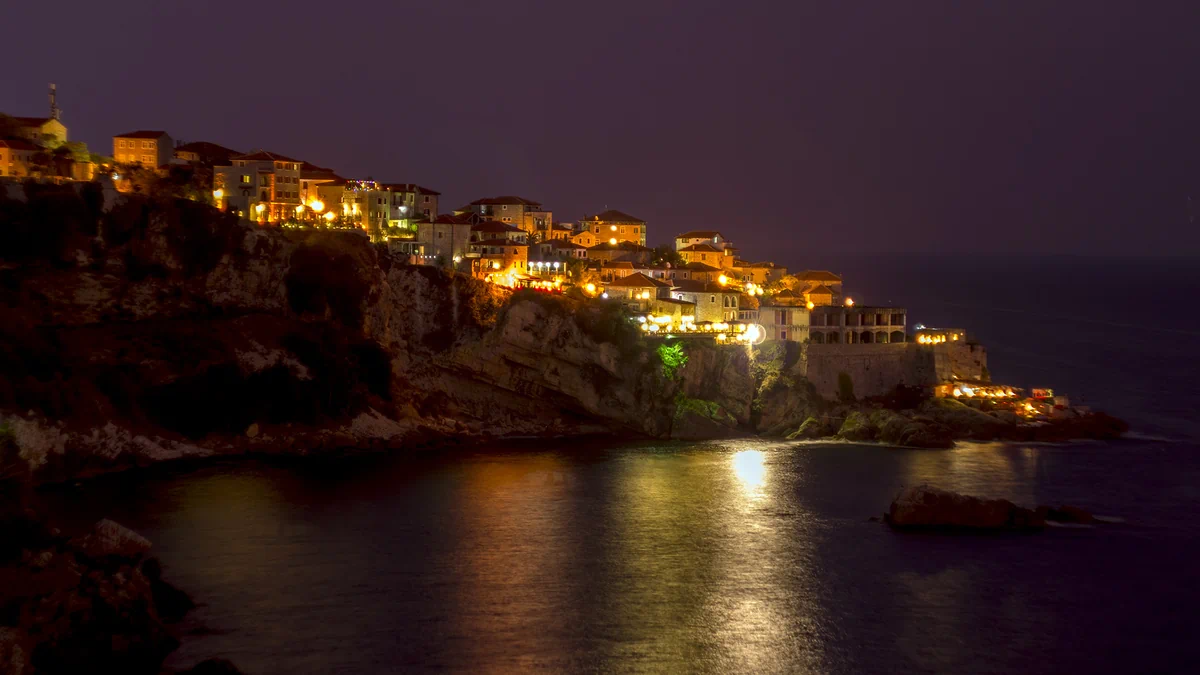 Ulcinj old town lit up at night on a rocky cliff above the Adriatic Sea, with warm lights reflecting on the dark water, Montenegro