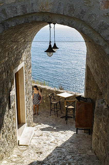 A woman standing under a stone arch in Ulcinj old town with a sea-view café terrace and the blue Adriatic in the background, Montenegro